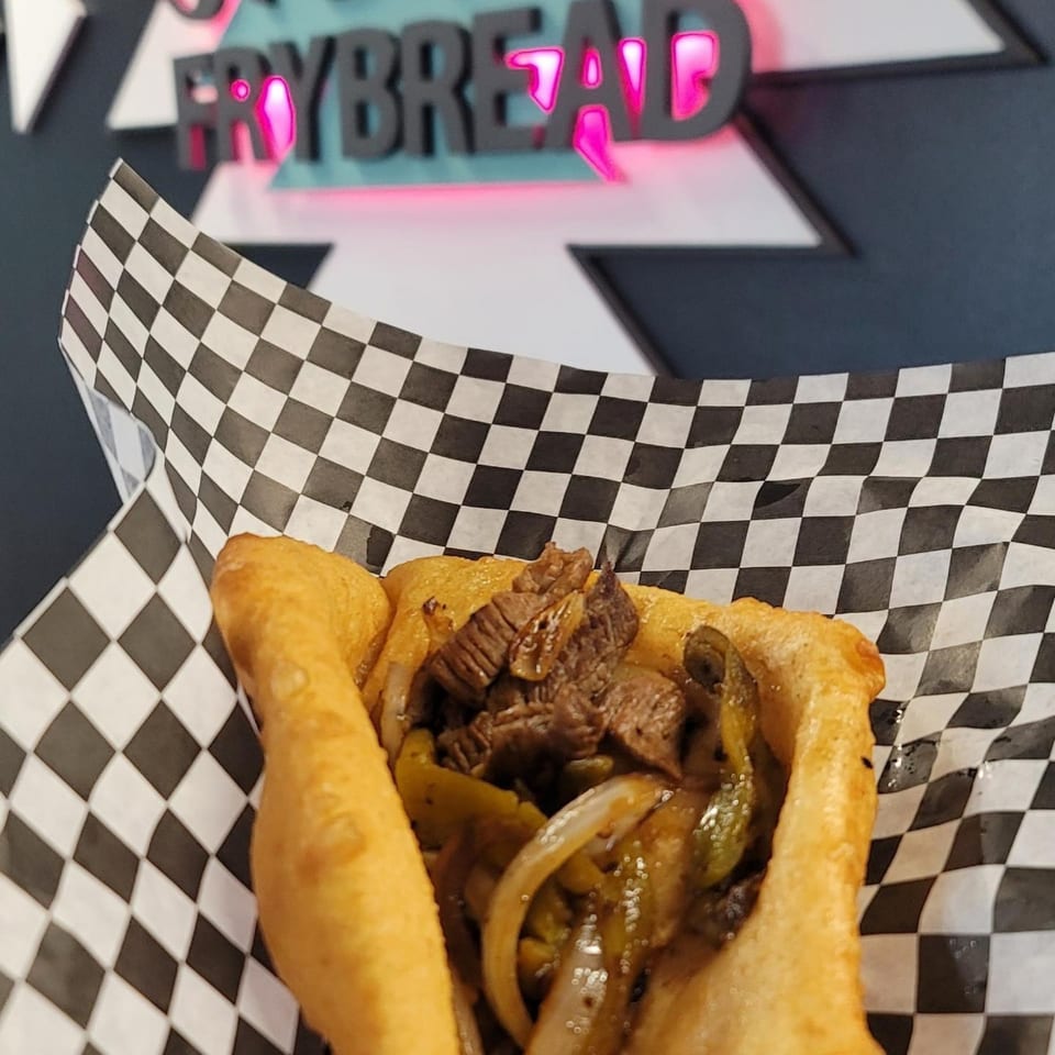 Hope's Frybread steak and onion frybread sandwich in checkered paper, with the restaurant sign in the background.