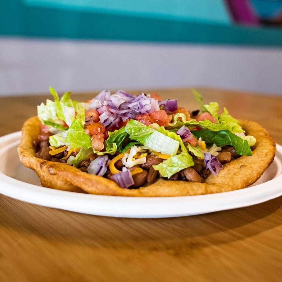 Hope's Frybread taco with beans, lettuce, tomatoes, and red onions on a white plate.